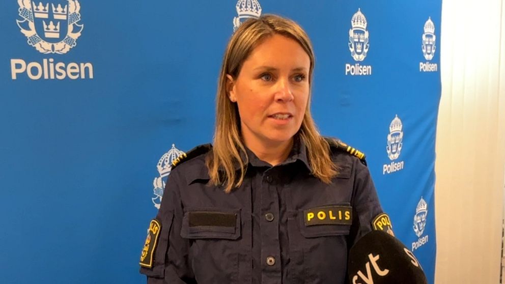 Polis i uniform intervjuas framför en blå bakgrund med polisens logotyp.