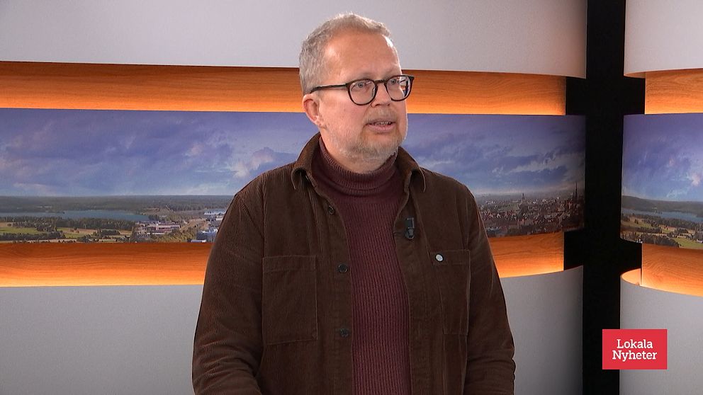 Personen är klädd i en mörkbrun jacka över en röd tröja och står framför en bakgrund med landskapsbilder, vilket tyder på en nyhetsstudio.