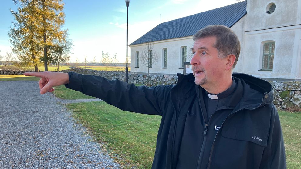 En kyrkoherde pekar med ena handen utanför Stenkvista kyrka. Det är en klar dag med ett par träd i bakgrunden och en lamppost syns i närheten. Gräset är grönt och välvårdat.