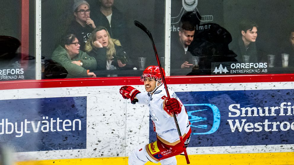 En hockeyspelare i röd och vit utrustning firar ett mål med utsträckt klubba och glädje. I bakgrunden syns åskådare vid en ishockeyrink.