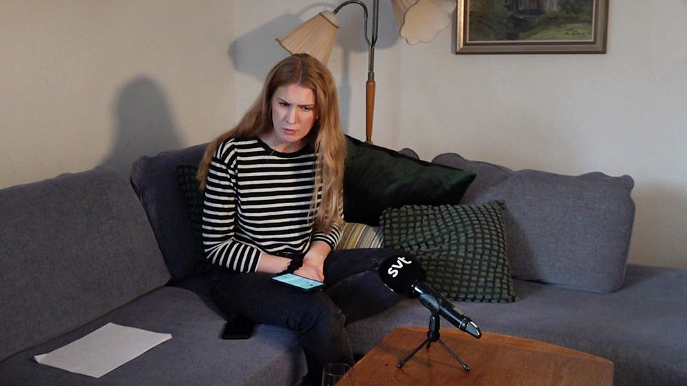 Kvinna sitter i soffa med mikrofon framför sig, anteckningar på soffan och mobil i handen.