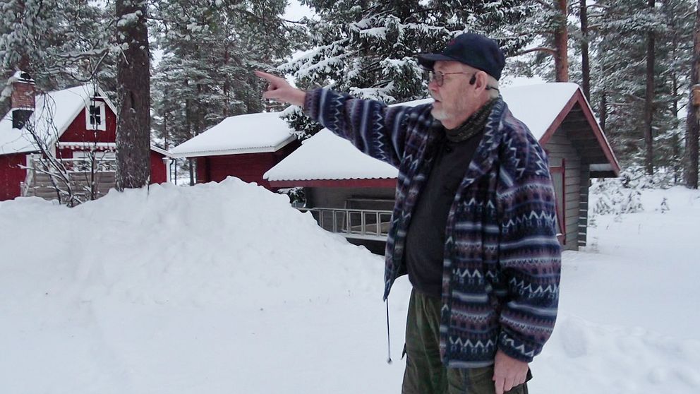 En person står i en snötäckt skog med en hand utsträckt och pekar mot en byggnad i bakgrunden. I närheten finns flera röda stugor omgivna av snötyngda träd. Det råder en kall och vintrig atmosfär.