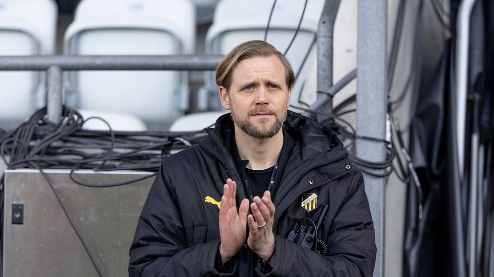 Person i svart sportjacka applåderar vid sidan av läktare med tomma stolar i bakgrunden.
