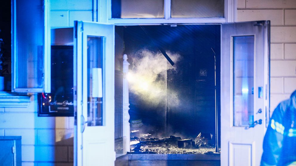Brandrök strömmar ur byggnadens öppna dörrar, blåljus reflekteras runtom.