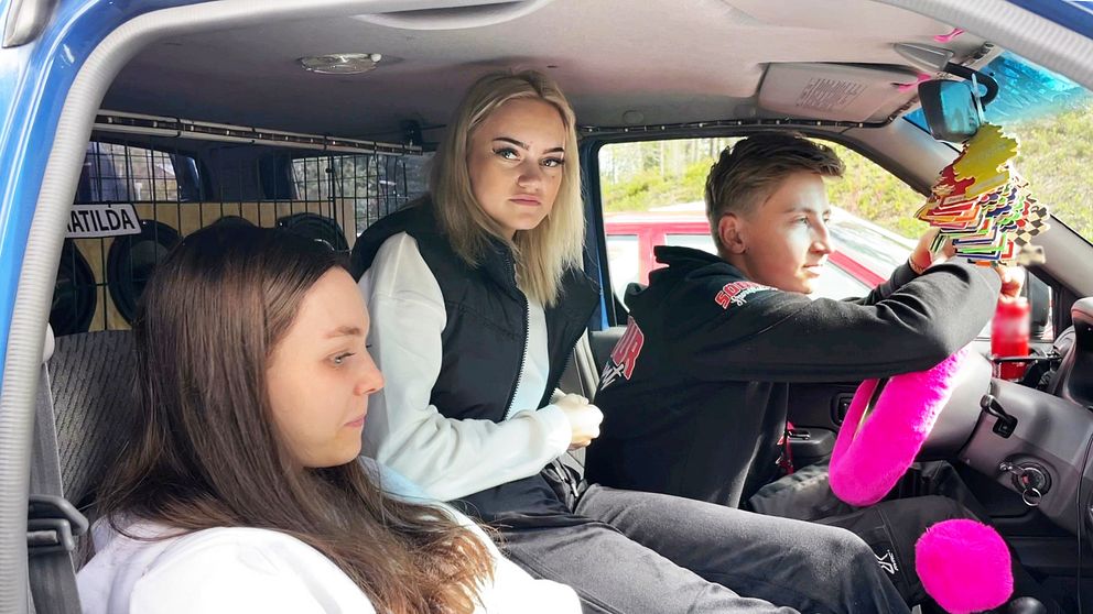 Tre ungdom sitter i en a-traktor – till vänster sitter en tjej med mörkt långt hår, i mitten av tjej med blont hår och vid ratten sitter en kortklippt kille. Det hänger wunderbaums, färggranna doftgranar, i fönstret runt backspegeln.