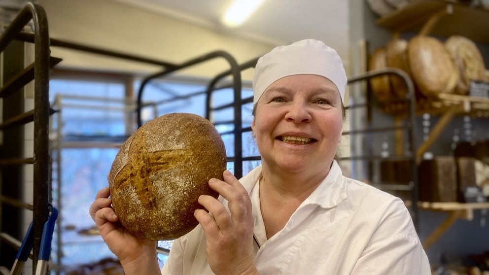 en glad kvinna i bagarekläder håller upp ett bröd. Tina Fernlund Järna
