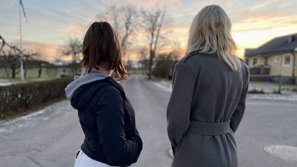 Två kvinnor står vända mot en väg vid solnedgången, med en ljus himmel och träd i bakgrunden. Den ena kvinnan bär en mörk jacka, medan den andra är klädd i en grå kappa. Scenen ger en känsla av reflektion och lugn.