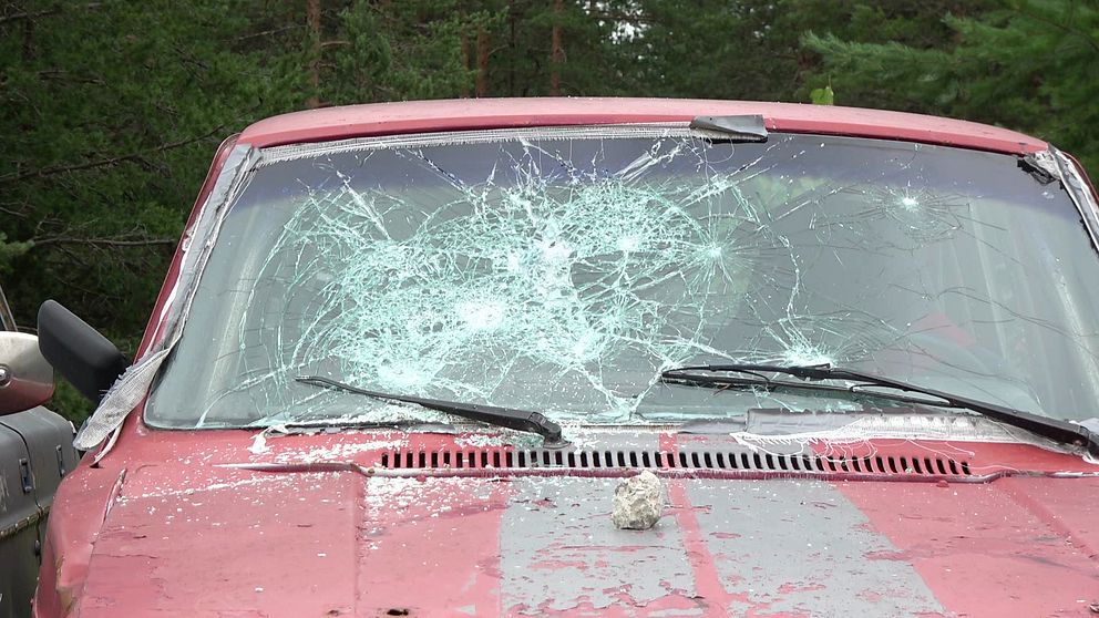 En röd Volvo är sönderslagen och en sten ligger på motorhuven under det krossade bilglaset.