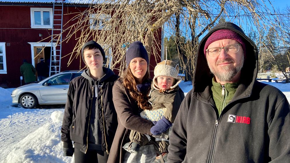 Tre vuxna och ett barn framför ett rött trähus med snö på backen.