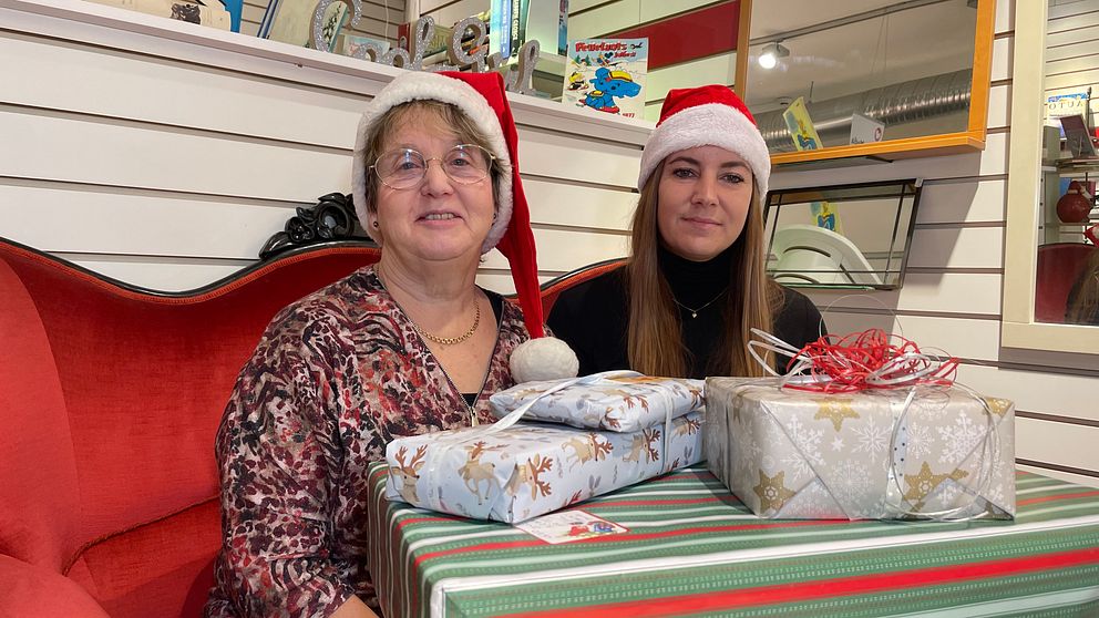 Två personer sitter på en röd soffa, klädda i juliga tomteluvor. Framför dem finns en julklappar med olika inslagningar, inklusive en med röd och vit design och en annan med silverpapper. Bakgrunden har hyllor med olika föremål, vilket ger en festlig atmosfär.