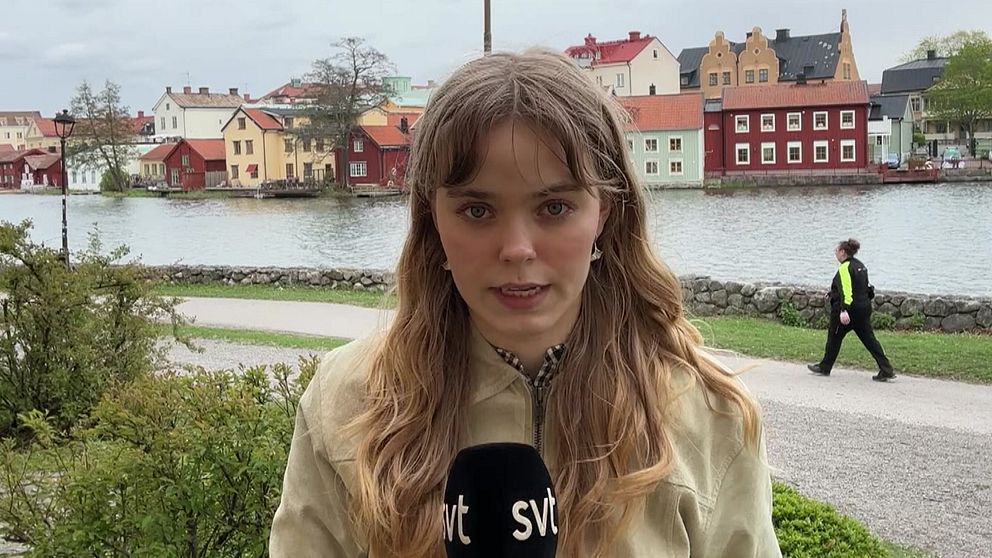 Reporter med SVT-mikrofon framför en å vid färgglada hus, en person promenerar i bakgrunden.