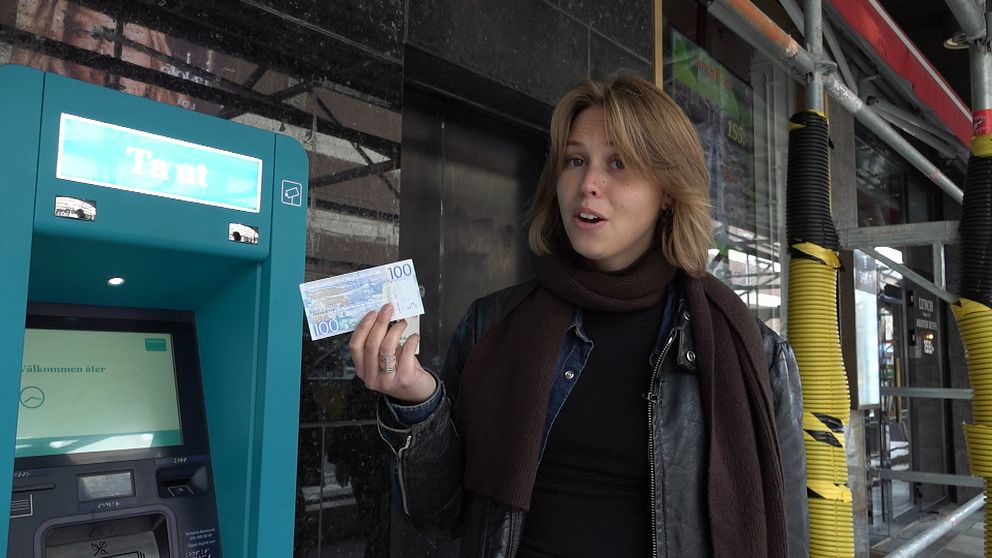 En person står vid en bankomat och håller upp en hundralapp. I bakgrunden syns en byggarbetsplats med rör och en glasruta.