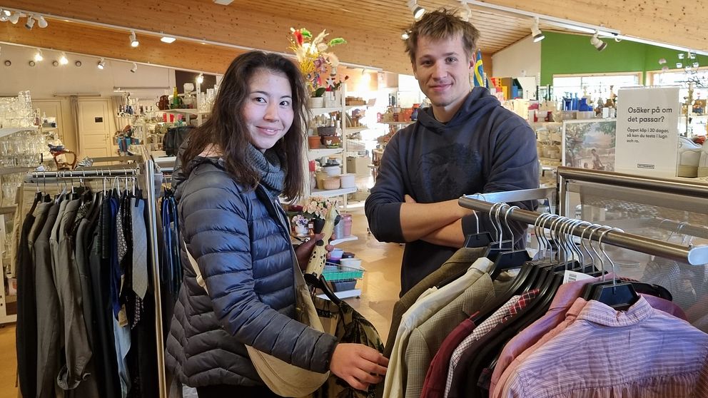 Två personer står i en butik med kläder och diverse inredning. Den första personen, klädd i en svart jacka, håller en väska och ser på kläder som hänger på en klädhängare. Den andra personen, i en blå tröja, står med armarna i kors och tittar på den andra personen. Butiken har ljusa väggar och en mängd olika varor syns i bakgrunden.