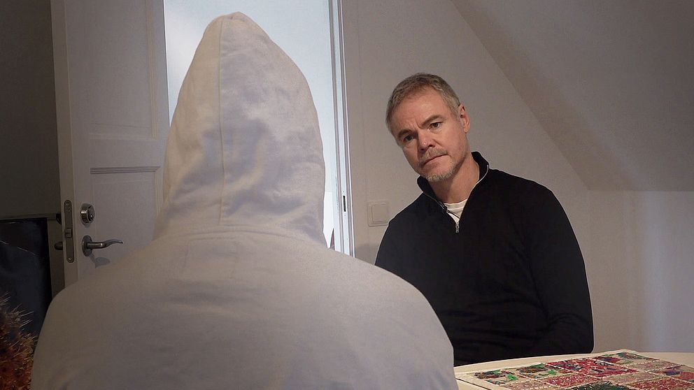 Två personer sitter mittemot varandra i ett inomhusmiljö. En person har på sig en vit huvtröja och är vänd bort från kameran, medan den andra personen, klädd i svart, sitter med fokus på den förra. Omkring dem finns en bordsyta med ett mönstrat föremål. Rummet har en modern inredning med ljusa väggar och en dörr i bakgrunden.