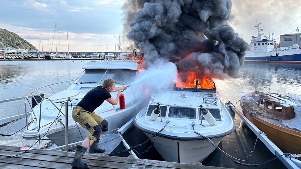 förtöjd motorbåt brinner kraftigt intill andra båtar, ihopkrupen man med brandsläckare försöker bekämpa lågorna från bryggan