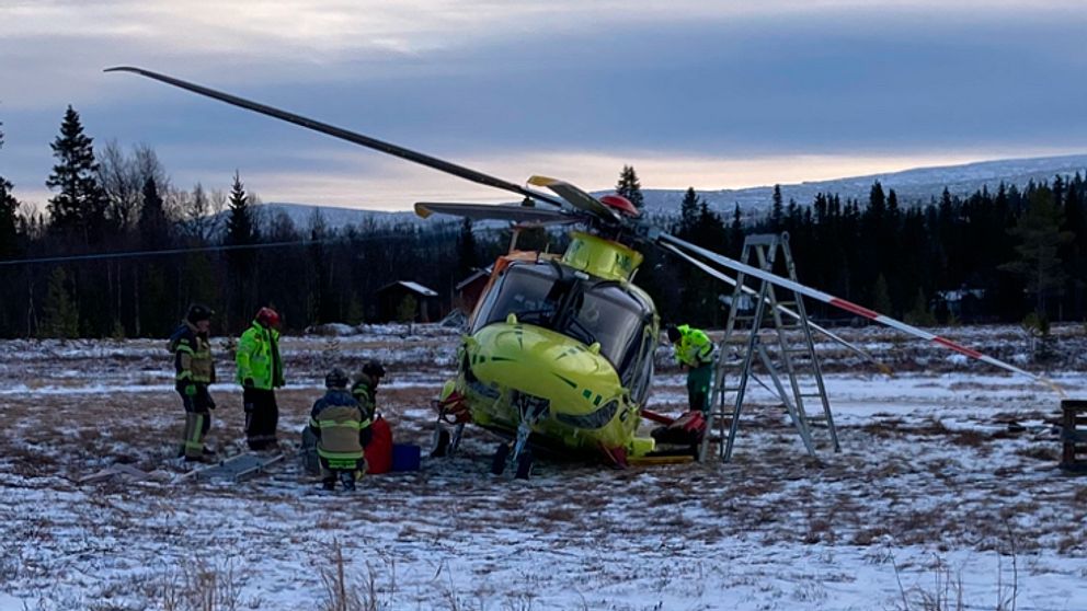Räddningspersonal arbetar runt en helikopter som har landat på en snötäckt äng. Helikoptern är grön och orange, och snön omger området. Några personer är på väg att hjälpa en patient i medvetslös tillstånd på marken. I bakgrunden syns träd och en fjärran bergskedja under en molnig himmel.