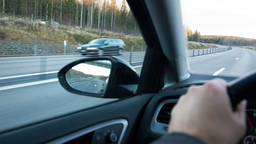 Vy från förarsätet i en bil som färdas på en motorväg, med en hand på ratten. En annan bil syns som kör i motsatt körfält. Tallskog och bar mark skymtas längs vägkanten.