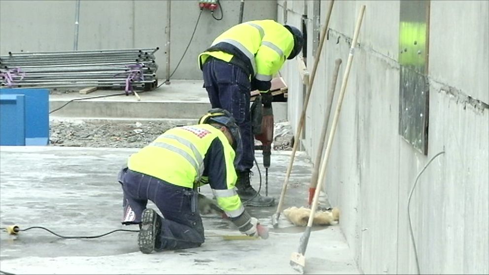 Två byggarbetare i reflexvästar arbetar på en byggplats. En av dem står och använder en jackhammer, medan den andra sitter på knä och mäter. I bakgrunden syns byggmaterial och en betongvägg.