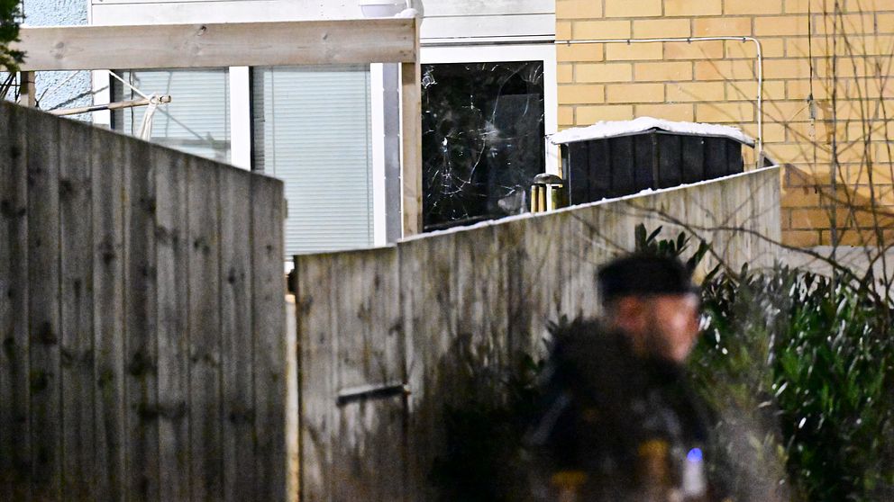 En man i uniform står framför en staketskärm med ett trasigt fönster i bakgrunden, vilket tyder på en eventuell incident. Området är svagt belyst och omges av buskar och byggnader.