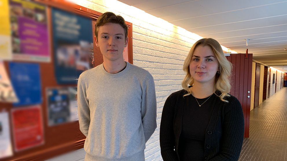Två elever står i en skolkorridor bredvid en svårläst anslagstavla