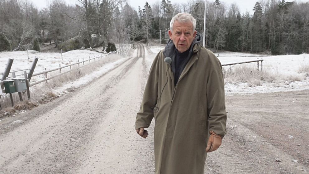 En person står på en grusväg i en snötäckt landskap, omgiven av träd. Vägen sträcker sig långt bort i bakgrunden, där den böjer sig. Det finns en postlåda intill vägkanten på vänster sida. Personen bär en lång, beige överrock.