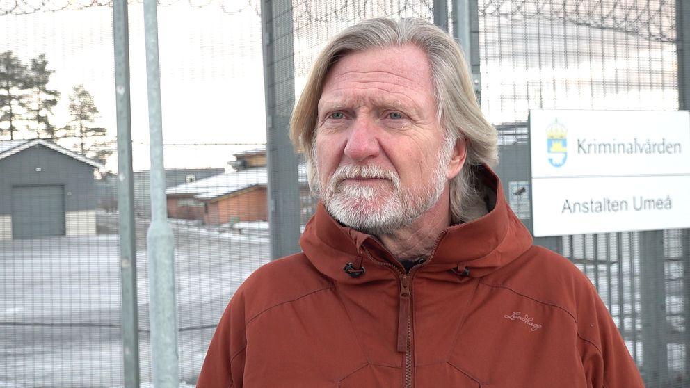 En person står framför ett stängsel på en anstalt, klädd i en röd jacka. I bakgrunden syns en byggnad samt en skylt med texten ”Kriminalvården Anstalten Umeå”. Det är en kall och klar dag, vilket framgår av den snöiga omgivningen.