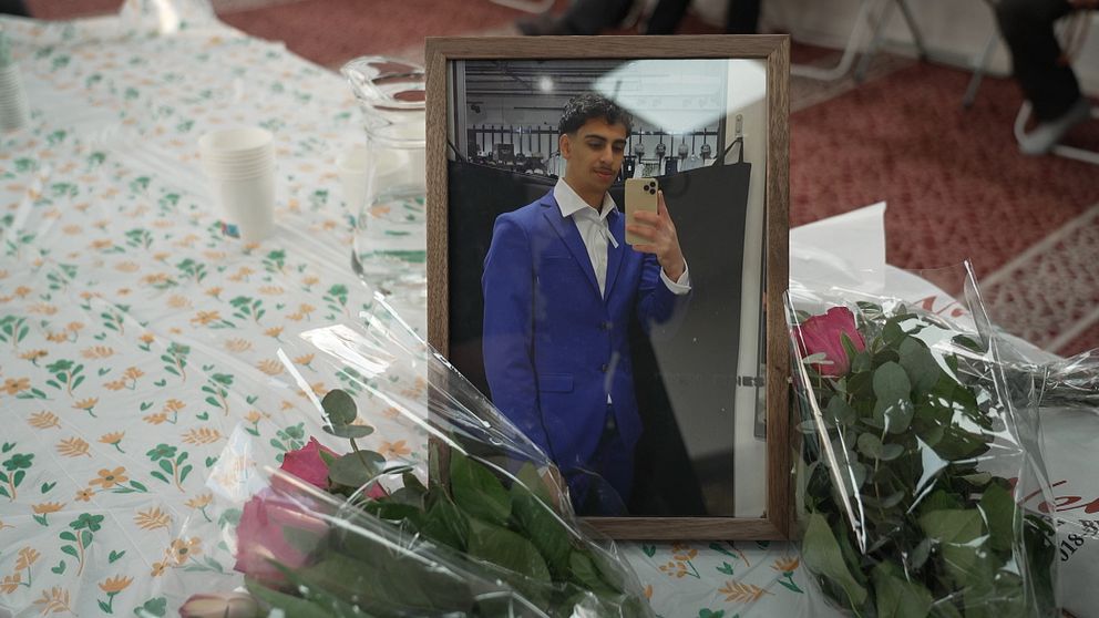 Inramat foto av en ung man, omgiven av rosor på ett blommigt bord.