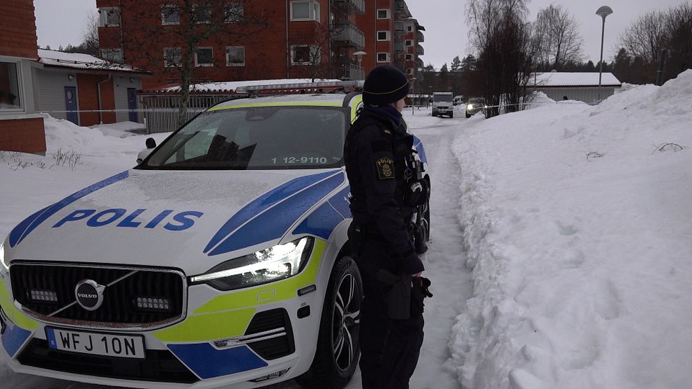 En polisbil står parkerad på en snöig gata framför en flerbostadsbyggnad. En polis i uniform står bredvid bilen och tittar bortåt längs gatan. Det är vinter och landskapet är täckt av snö.