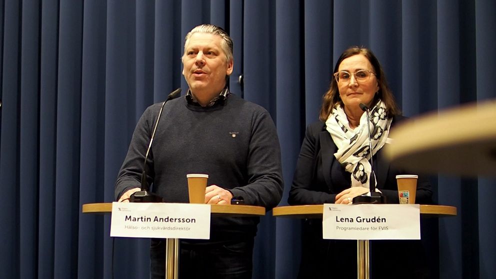 Två personer står vid podier under en presskonferens med namnskyltar framför sig. Den ena personen är klädd i en mörk tröja och den andra har en sjal runt halsen. Båda har pappersmuggar framför sig och det finns en blå bakgrund av tyg bakom dem.
