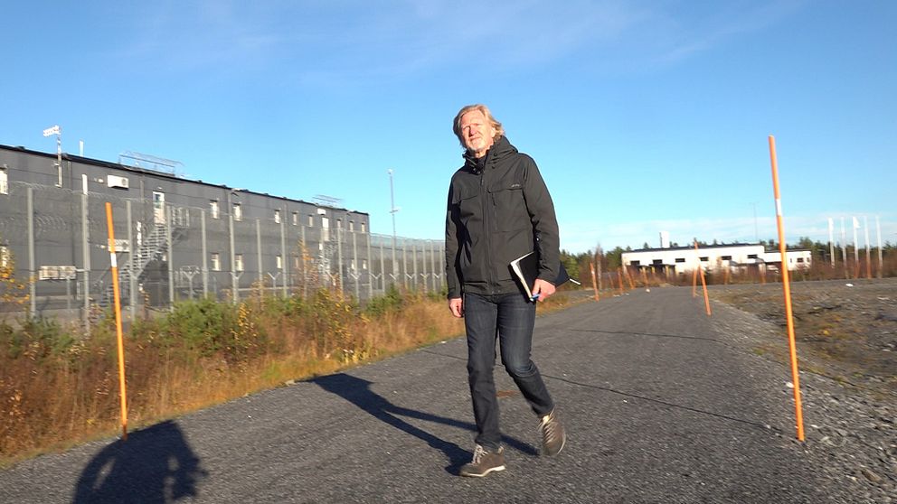 En person går längs en asfaltväg som leder förbi en inhägnad byggnad. Omgivningen är grön och solig, med en klar blå himmel. Längs vägkanten står orangea markörer.