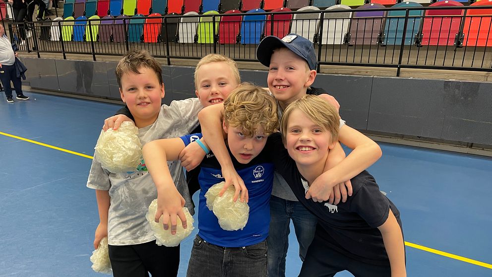 Fyra ungdomar står eniga med vita pom-poms inomhus framför läktarsäten.