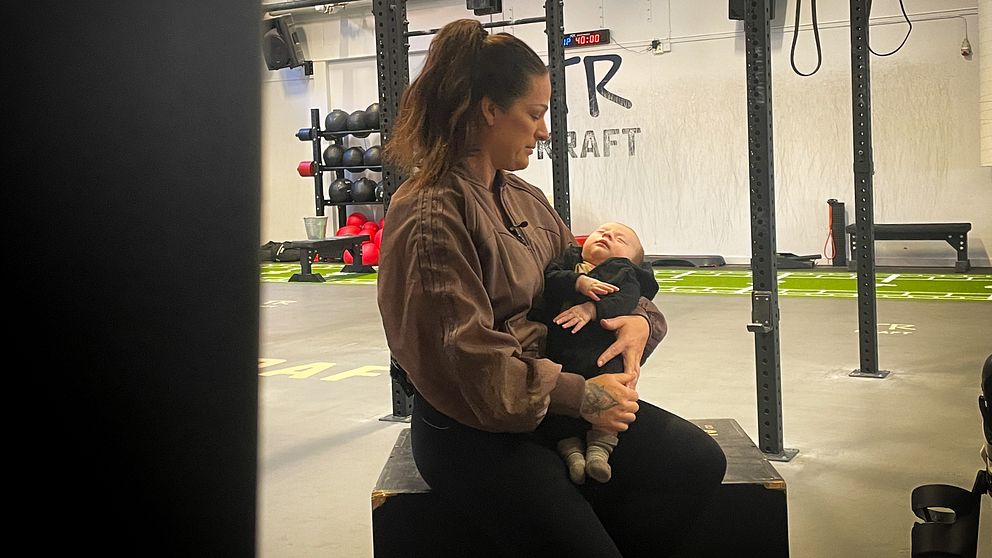 Kvinna sitter i ett gym med en bebis i famnen.