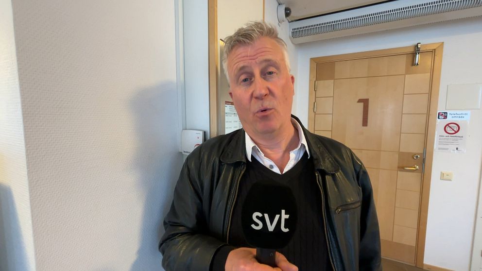 Person med grått hår håller en SVT-mikrofon i en korridor utanför en dörr märkt med en etta.