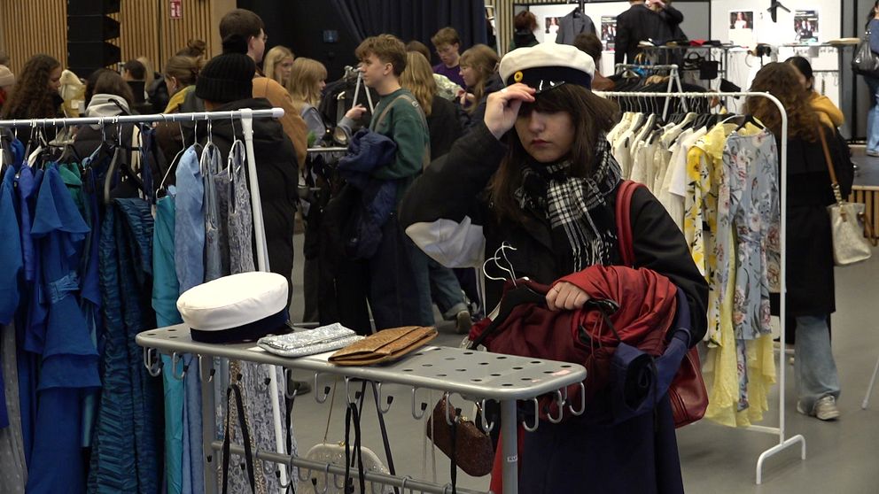 En tjej testar en studentmössa i förgrunden. Kläder i olika färger och mönster hänger på klädställningar runtomkring, och lokalen är fylld med människor.