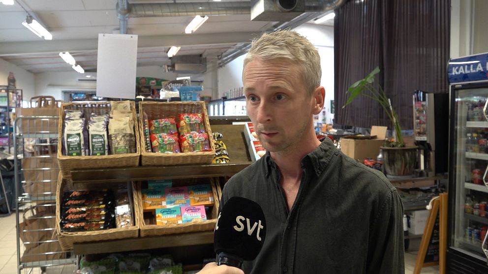 En man med blont hår och skjorta står i vad som ser ut att vara en liten matbutik.