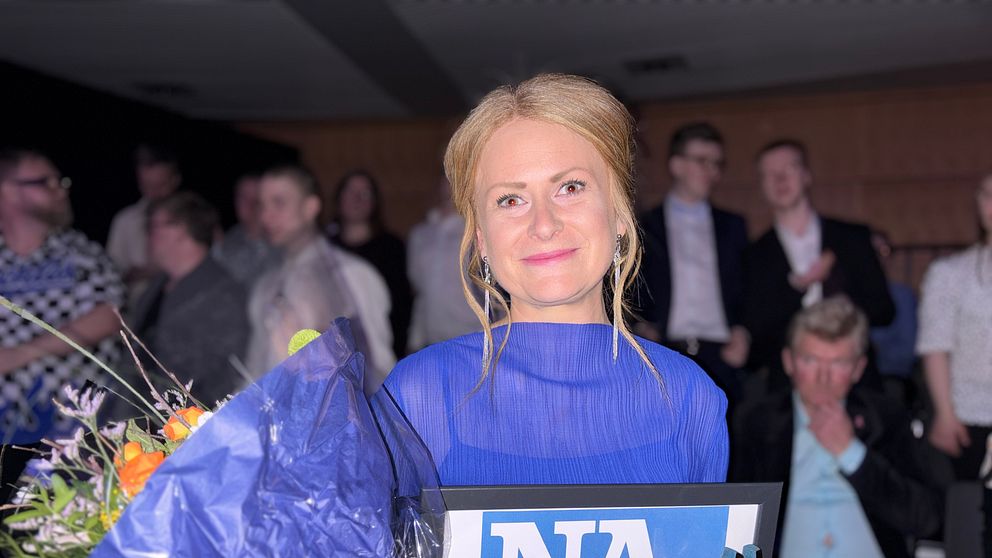 Stina Öst i blå festklänning håller blombukett och plakett med texten ”NA” och ”JAG ÄR VINNARE” inför publik.