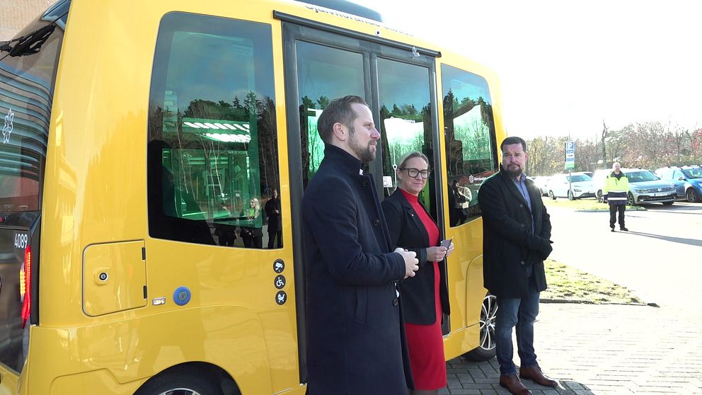 Tre personer står framför en gul självkörande buss. Två av dem är klädda i mörka rockar, medan den tredje bär en röd klänning. Bussen är parkerad på en asfaltväg, med en ljus bakgrund av träd och byggnader.