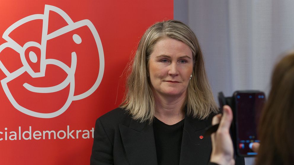 En person i mörk kavaj står framför en röd bakgrund med Socialdemokraternas logotyp, medan en annan person tar ett foto med en mobiltelefon.