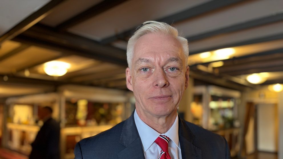 Man i kostym och röd slips står i receptionen. En annan person syns i bakgrunden.
