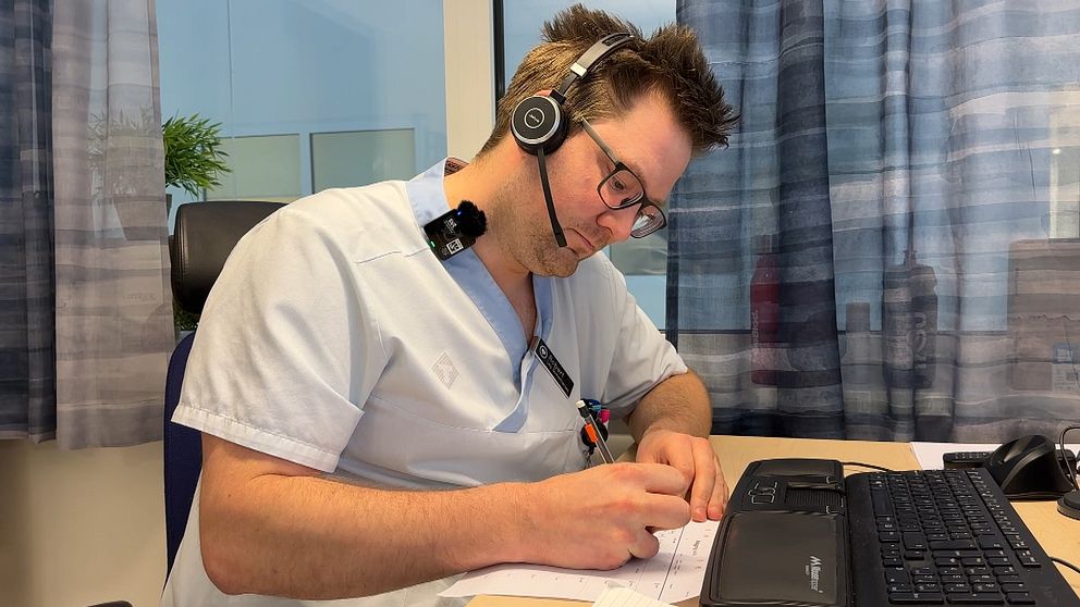 En sjuksköterskeklädd man sitter vid ett skrivbord och skriver för hand. På huvudet har han ett headset.