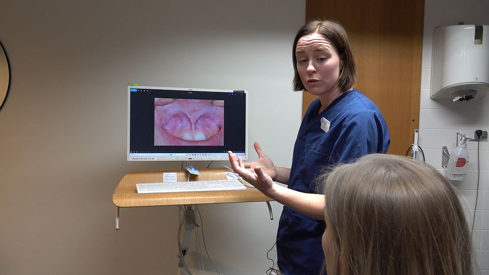 Tandläkaren Emma Wider står framför en datorskärm som visar en närbild av en patients mun med tänder. Personen gestikulerar medan en annan person, som deltar i diskussionen, sitter vänd mot dem. Rummet ser ut att vara en tandvårdsklinik med nödvändig utrustning i bakgrunden.