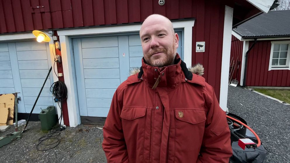 En person i en röd jacka står framför en röd byggnad med blå garageportar. På vänster sida finns en lampa som lyser, och i bakgrunden syns en uppfart med grus samt en del av en staket inramande området.