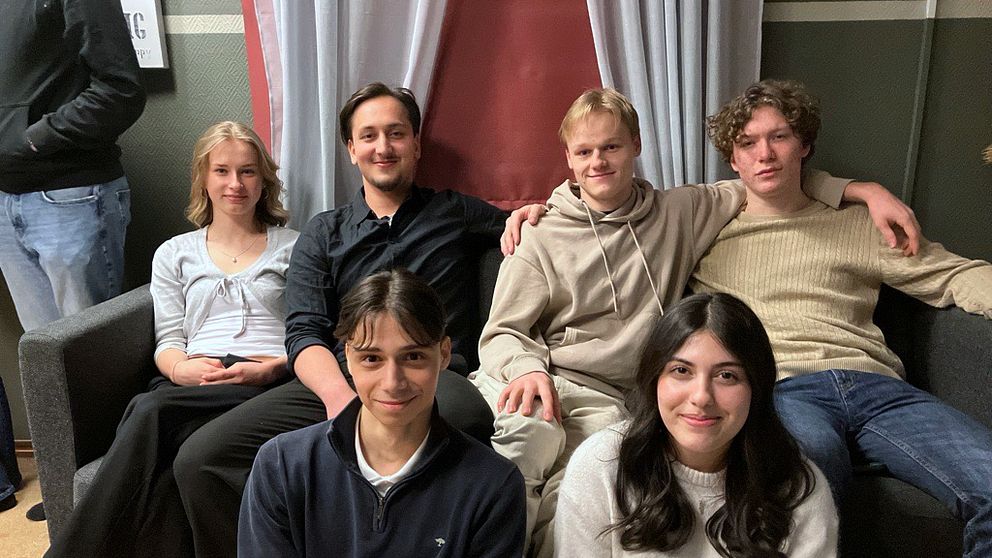 En grupp av sex personer sitter och står tillsammans i ett vardagsrumsliknande utrymme. Fyra personer sitter tätt ihop i soffan, medan två personer sitter på golvet framför dem. Det finns vita gardiner och en röd vägg i bakgrunden.