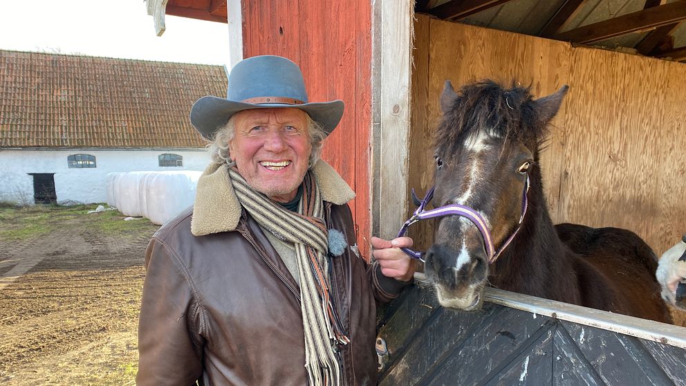 En person i läderjacka och scarf står vid en hage och interagerar med en svart häst som sträcker sig fram genom stallets öppning. I bakgrunden syns en lada och andra delar av gården.