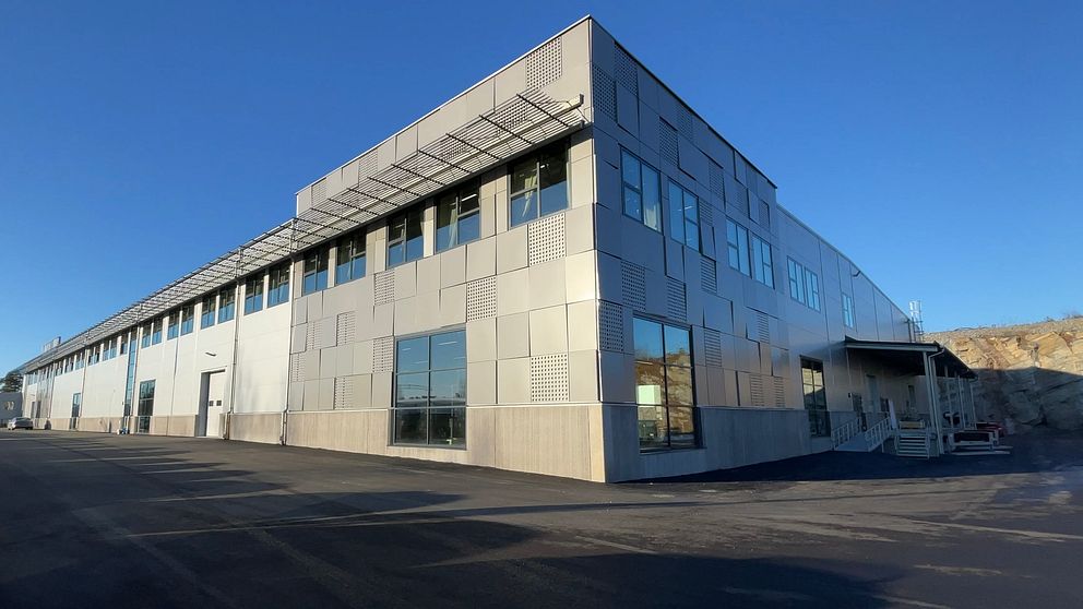 Industribyggnad i solljus med moderna fönster och platt tak, belägen på en asfalterad yta under en klarblå himmel.