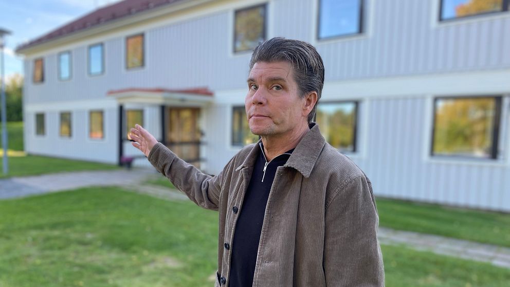 Mattias Fjellström ny chef på Sis ungdoms Vemyra står utomhus och gestikulerar med en arm, framför en byggnad med flera fönster. Området runt dem är grönt med gräsmatta och en stenlagd gång. Byggnaden har en ljus färg och ett rött tak, vilket ger en kontrast till den omgivande naturen.
