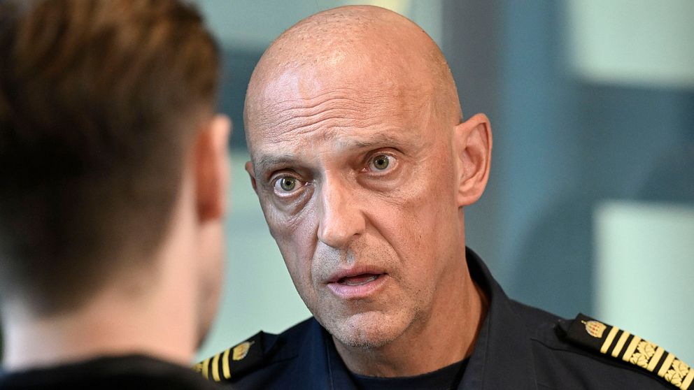 Två personer samtalar, en i polisuniform och en med ryggsäck, inomhus vid grönt draperi.