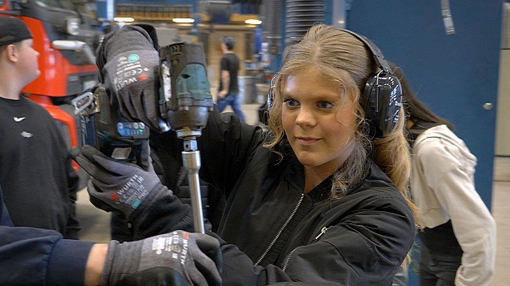 tjej arbetar med mutterdragare i verkstad, flera elever syns i bakgrunden