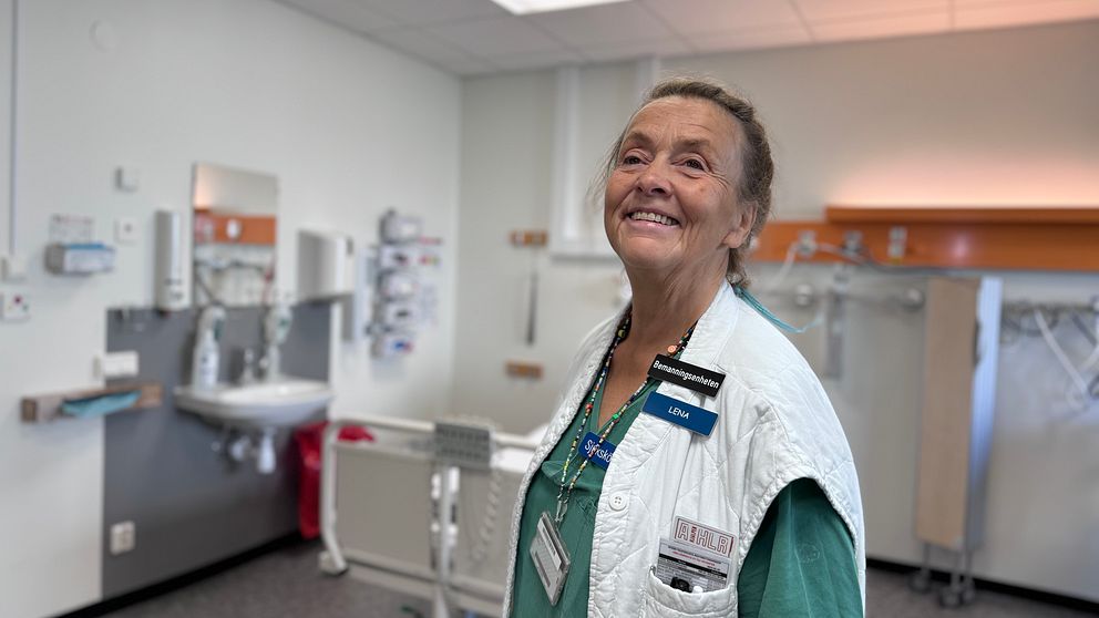 Medarbetare i sjukhusmiljö, klädd i vit väst och gröna kläder, står i ett rum med medicinska apparater och utrustning i bakgrunden. Hon ler mot kameran. Rummet är ljust och modernt inrett, med ett handfat och sjukhuslådor synliga.