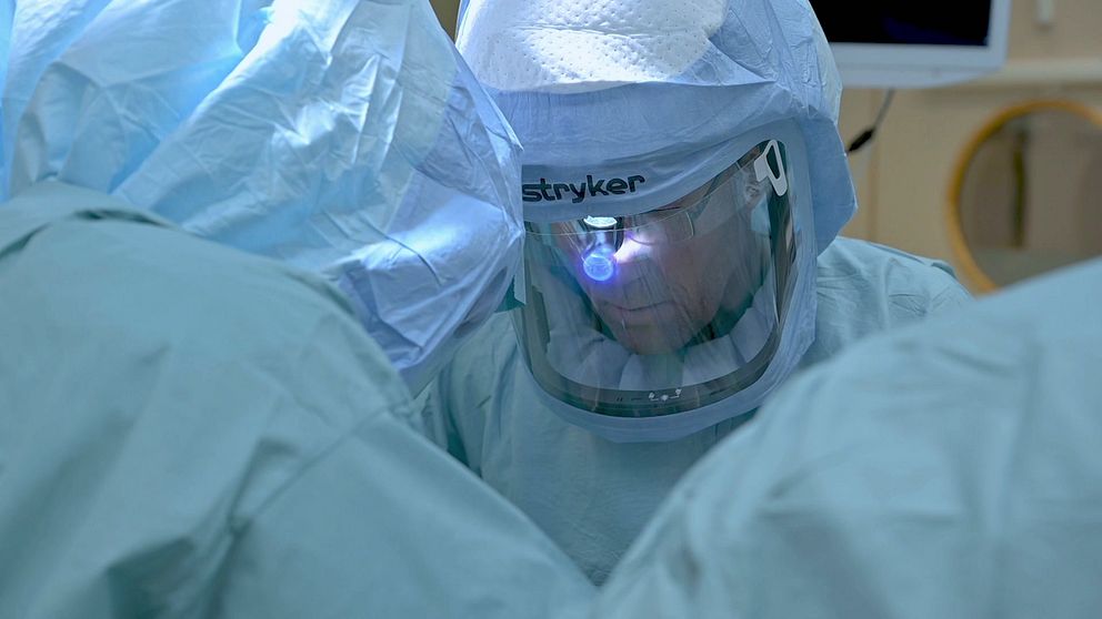Opererande kirurg i skyddskläder med visir och pannlampa.